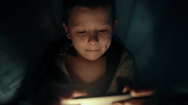 Pequeño chico debajo de la manta está jugando un juego en su teléfono inteligente por la noche — Vídeo de stock