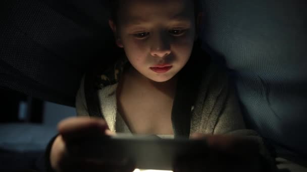 Pequeño chico debajo de la manta está jugando un juego en su teléfono inteligente por la noche — Vídeos de Stock