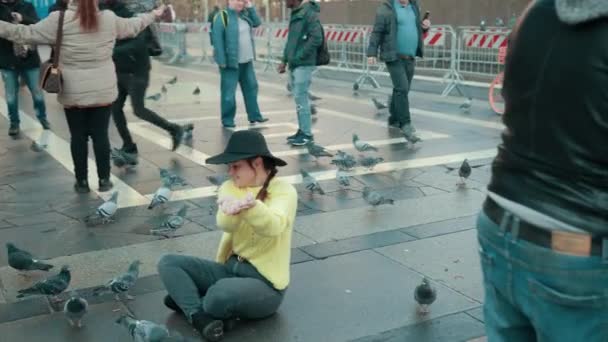 Jeune fille en pull jaune avec chapeau s'assoit sur la tuile et nourrit les pigeons avec les mains . — Video