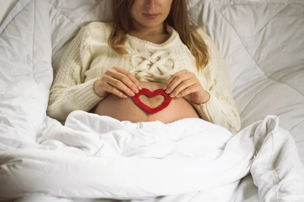 Jovem grávida na cama de barriga segura o coração vermelho de — Fotografia de Stock