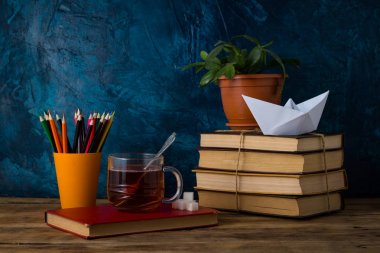 Kitap, bir fincan sıcak çay, bir kağıt tekne, renkli kalemler yığını