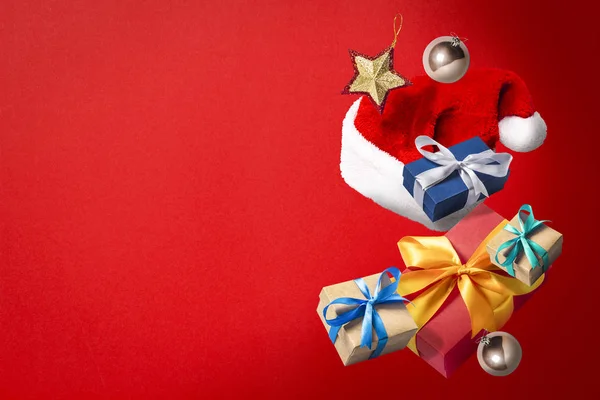 Létající Santa Claus klobouk, dárkové krabice a vánoční ozdoby na červeném pozadí. Koncept svátků, Vánoce. Levitace. Věci ve vzduchu. Byt ležel, horní pohled — Stock fotografie