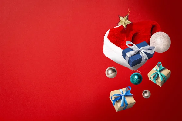 Chapeau de Père Noël volant, boîte cadeau et décorations de Noël sur un fond rouge. Concept de vacances, Noël. Lévitation. Des objets en l'air. Couché plat, vue du dessus — Photo