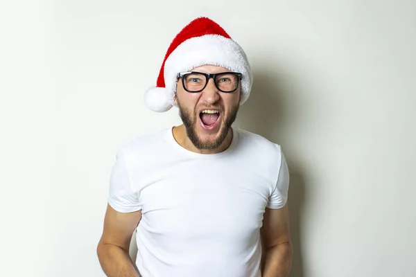 Ung man i glasögon och en tomte hatt skriker på en vit bakgrund. Julkoncept. Gesture seger, super, skål, glädje — Stockfoto