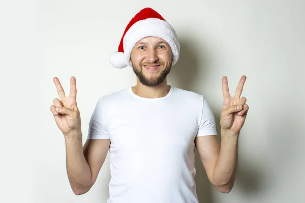 A young man in a Santa Claus hat makes a gesture with his hands on a white background. Christmas concept. Gesture of victory, super — Stock Photo, Image