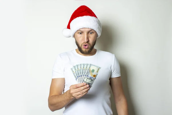 Ein junger Mann mit überraschtem Gesicht und Weihnachtsmann-Mütze hält auf weißem Hintergrund ein Bündel Geld in den Händen. Weihnachtsverkaufskonzept, Schwarzer Freitag, Rabatt, reich, gewinnen, wetten — Stockfoto