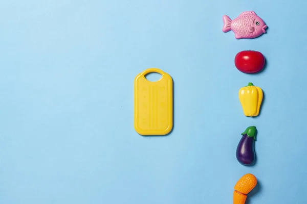Yellow cutting board and toys in the form of food on a blue background. Concept Artificial food, proper nutrition, diet, convenience foods. Flat lay, top view