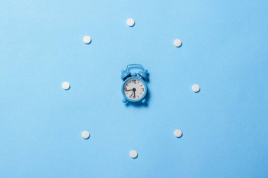 Haplar yuvarlak arama şeklinde ve mavi arka planda bir çalar saat şeklinde dizilmişti. Uykusuzluk konsepti, rejime göre haplar, vitaminler. Düz yatış, üst görünüm