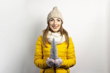 Sıcak şapkalı ve eldivenli genç bir kadın gülümsüyor ve arka planda dekoratif bir Noel ağacı tutuyor. Kış kavramı, kış tatili, Noel, yeni yıl. Komik ruh hali, duygular, sürpriz