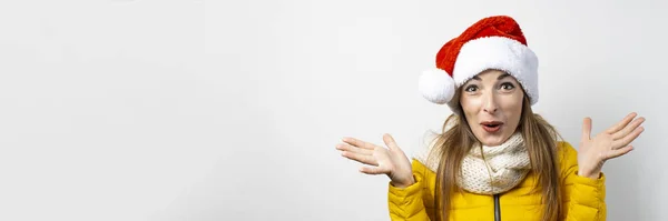 Mujer joven con sombrero de Santa Claus extiende sus brazos sobre un fondo claro. Concepto de invierno, vacaciones de invierno, Navidad, Año Nuevo. Humor divertido, emociones, sorpresa, shock. Banner — Foto de Stock