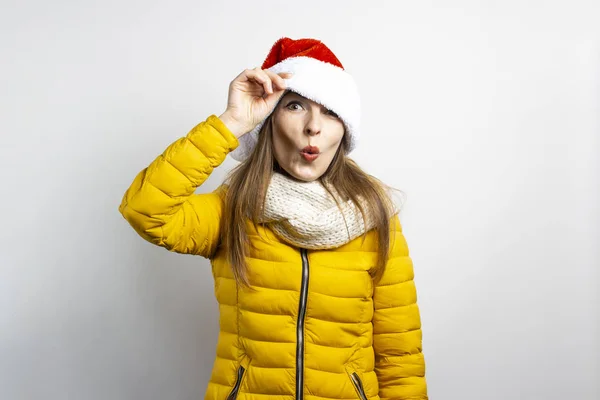 Mujer joven mira desde debajo de los sombreros de Santa Claus con los ojos puestos en un fondo claro. Concepto de invierno, vacaciones de invierno, Navidad, Año Nuevo. Humor divertido, emociones, sorpresa, shock — Foto de Stock