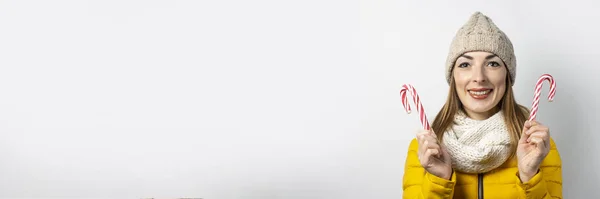 Mujer joven con un sombrero y una chaqueta de plumón sostiene un bastón de caramelo sobre un fondo claro. Forma de corazón. Concepto invierno, navidad, vacaciones de invierno. Banner — Foto de Stock