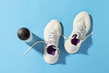 Beyaz spor ayakkabılar ve sabah güneşinin altında mavi bir arka planda bir şişe su. Koşu, koşma, spor, çapraz forma, sıkı çalışma. Sabah koşusu. Pankart. Düz yatış, üst görünüm