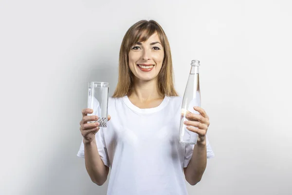 Une jeune femme tient une bouteille et un verre avec de l'eau claire sur un fond clair. Bannière. Concept de soif, chaleur, santé et soins de beauté, équilibre hydrique — Photo