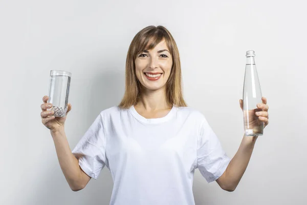 Une jeune femme tient une bouteille et un verre avec de l'eau claire sur un fond clair. Bannière. Concept de soif, chaleur, santé et soins de beauté, équilibre hydrique — Photo