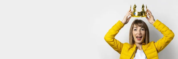 Young woman in a yellow jacket puts a golden crown on her head on a light background. Emotion laughter, surprise, kiss. Concept of a queen, luck, the best, win, victory. Banner — Stock Photo, Image