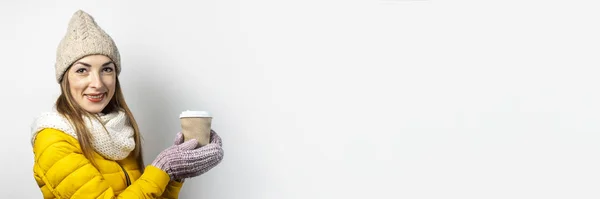 Mujer joven con una chaqueta amarilla y sombrero sostiene un vaso de café o té sobre un fondo claro. Risa de emoción, sorpresa, beso. Concepto invierno, otoño, cafetería. Banner — Foto de Stock