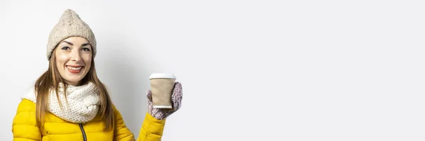 Junge Frau in gelber Jacke und Hut hält ein Glas Kaffee oder Tee vor hellem Hintergrund. Emotionales Lachen, Überraschung, Kuss. Konzeptwinter, Herbst, Kaffeehaus. Banner — Stockfoto
