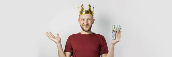 Young man in a golden crown with a surprised face holds money in his hands on a light background. Concept is king, luck, gain, rich, dream, goal, aspiration, bet. Banner — Stock Photo, Image