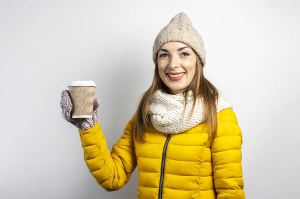 Young woman in a yellow jacket and hat holds a glass of coffee or tea on a light background. Emotion laughter, surprise, kiss. Concept winter, autumn, coffee house