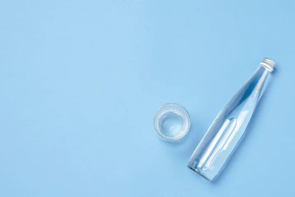 Μπουκαλάκι γυάλινο και γυάλινο με διαυγές νερό σε μπλε φόντο. Έννοια της υγείας και της ομορφιάς, ισορροπία νερού, δίψα, ζέστη, καλοκαίρι. Επίπεδο lay, πάνω όψη — Φωτογραφία Αρχείου