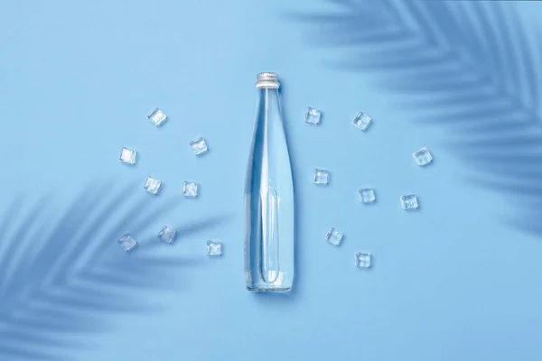 Skleněná láhev s čirou vodou na modrém pozadí s kostkami ledu. Koncept zdraví a krásy, vodní bilance, žízeň, teplo, léto. Plocha, výhled shora. Nápis — Stock fotografie