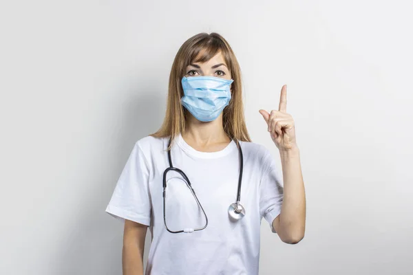 Ung kvinna i medicinsk mask och stetoskop pekar finger på en ljus bakgrund. Banner. Begreppsläkare, medicin på hög nivå, vaccination, läkarbesök — Stockfoto