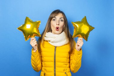 Sarı ceketli, şaşırmış yüzlü genç bir kadın mavi arka planda yıldız balonları tutuyor. Kış ve sonbahar kavramı, parti, tatil. Pankart