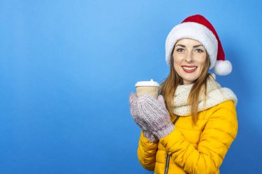 Gülümseyen ve Noel Baba şapkalı genç bir kadın elinde kahve, çay ya da mavi arka planda sıcak şarap olan bir kağıt bardak tutuyor. Tatil kavramı, Noel, yeni yıl, sürpriz, şok. Pankart