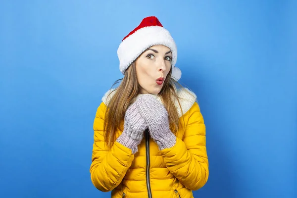 Young woman with a surprised face in a yellow jacket and a hat of Santa Claus on a blue background. Concept of the winter holidays, Christmas, New Year, surprise, shock. Banner