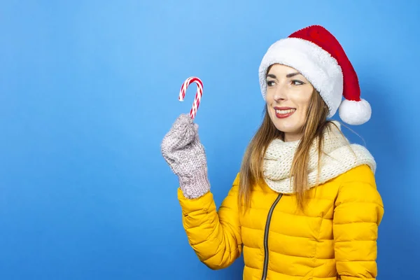 Mujer joven con una sonrisa en una chaqueta amarilla y un sombrero de Santa Claus sostiene un bastón de caramelo sobre un fondo azul. Concepto de las vacaciones de invierno, Navidad, Año Nuevo, sorpresa, shock. Banner — Foto de Stock