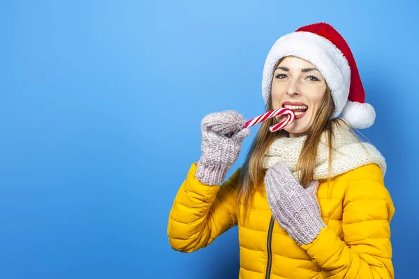 Mujer joven con una sonrisa en una chaqueta amarilla y un sombrero de Santa Claus sostiene un bastón de caramelo sobre un fondo azul. Concepto de las vacaciones de invierno, Navidad, Año Nuevo, sorpresa, shock. Banner — Foto de Stock