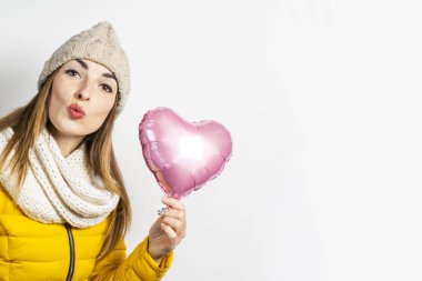 Sarı ceketli ve şapkalı genç bir kadın kalp şeklinde bir hava balonu tutuyor ve arka planda hafif bir öpücük yolluyor. Sevgililer günü konsepti, hediye, tatil, sevgili. Pankart
