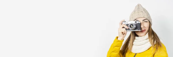 Jovem mulher de casaco amarelo e chapéu segura uma câmera em suas mãos e tira uma foto em um fundo claro. Conceito de beleza e moda, inverno, outono, tecnologia. Banner — Fotografia de Stock