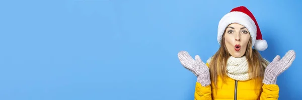 Mujer joven con una cara sorprendida en una chaqueta amarilla y un sombrero de Santa Claus sobre un fondo azul. Concepto de las vacaciones de invierno, Navidad, Año Nuevo, sorpresa, shock. Banner — Foto de Stock