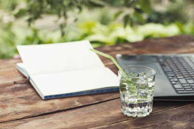 Laptop, açık günlüğü ve bir bardak buz küpleri ve nane ile ferahlatıcı bir içki ile ahşap bir masaya bırakır. Doğada, freelancing, tatilde iş iş kavramı