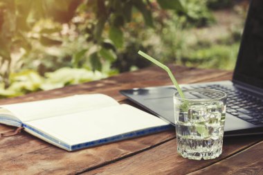 Laptop, açık günlüğü ve bir bardak buz küpleri ve nane ile ferahlatıcı bir içki ile ahşap bir masaya bırakır. Doğada, freelancing, tatilde iş iş kavramı