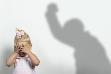 Little child covers his face with his hands on a light background with the shadow of an attacking man. Concept of fear, fear, domestic violence, the game of locks