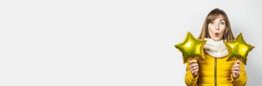 Sarı ceketli, şaşkın yüzlü ve beyaz arka planda yıldız balonları tutan bir kadın. Tatil, kutlama, parti, yeni yıl kavramı. Pankart
