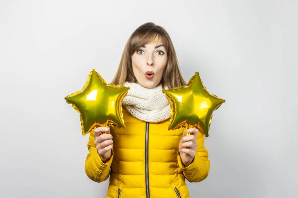 Mujer con una cara sorprendida con una chaqueta amarilla y una bufanda que sostiene globos de aire estrellados sobre un fondo blanco. El concepto de la fiesta, la fiesta, la fiesta, un nuevo año. Banner — Foto de Stock