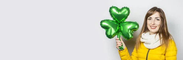 Woman with a smile in a yellow jacket and a scarf holds a clover balloon on a white background. Concept of a holiday, celebration, party, St. Patrick\'s Day, Ireland. Banner