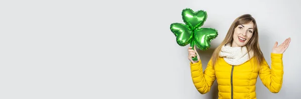 Woman with a smile in a yellow jacket and a scarf holds a clover balloon on a white background. Concept of a holiday, celebration, party, St. Patrick's Day, Ireland. Banner — Stock Photo, Image