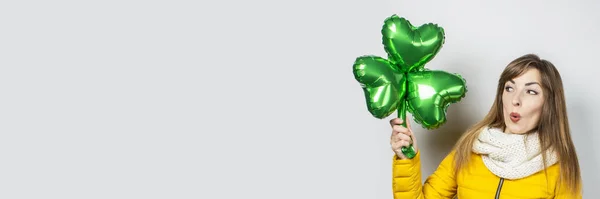 Woman with surprise in a yellow jacket and scarf holds a clover balloon on a white background. Concept of a holiday, celebration, party, St. Patrick's Day, Ireland. Banner — Stock Photo, Image