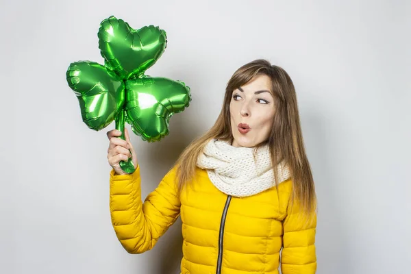 Woman with surprise in a yellow jacket and scarf holds a clover balloon on a white background. Concept of a holiday, celebration, party, St. Patrick's Day, Ireland. Banner — Stock Photo, Image