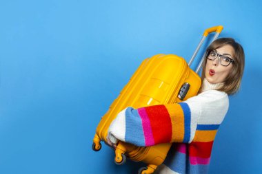 Şaşırmış yüzlü genç bir kadın mavi arka planda sarı bir bavul tutuyor. Konsept seyahat, seyahat, tatil. Duygusallık. Pankart