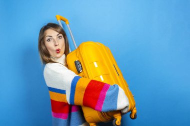 Şaşırmış yüzlü genç bir kadın mavi arka planda sarı bir bavul tutuyor. Konsept seyahat, seyahat, tatil. Duygusallık. Pankart