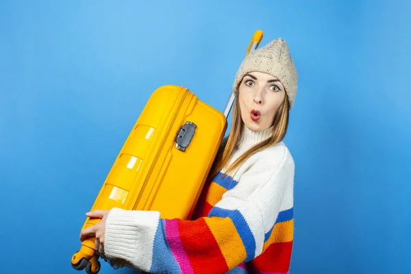 Young woman in a hat with a surprised face holds a yellow suitcase on a blue background. Concept of travel, trip, vacation. Emotional. Banner — Stock Photo, Image