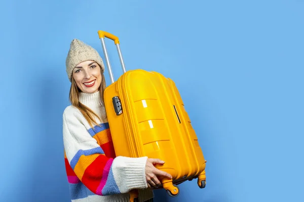 Eine junge Frau mit Hut hält lächelnd einen gelben Koffer vor blauem Hintergrund. Konzept der Reise, Reise, Urlaub. Frühling, Winter. Banner — Stockfoto