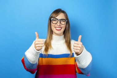 Gözlüklü ve örgü kazaklı genç bir kız düz ve gülümsüyor, mutlu, elleriyle, klasıyla, mavi arka planda süper bir jest yapıyor. Pankart.