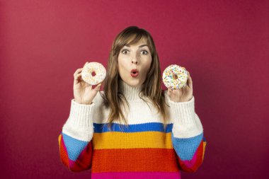 Örgü kazaklı genç bir kız elinde beyaz kremalı ve çok renkli tozlu iki donut tutuyor, şarap rengi bir arka planda. Pankart.